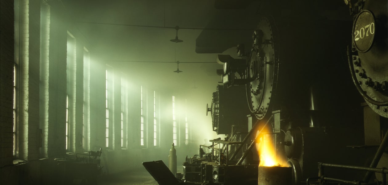 Steam locomotives standing in a roundhouse, lit by a row of tall windows through the smoke, with a fire burning beside the nearest engine